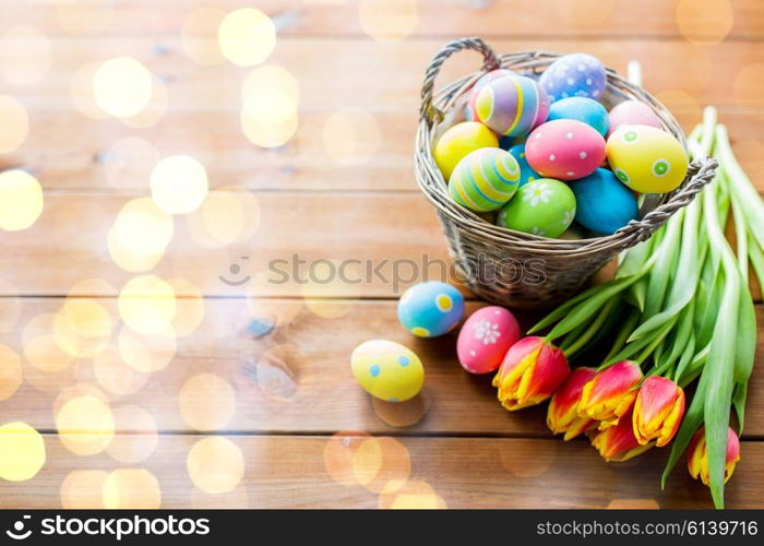 easter, holidays, tradition and object concept - close up of colored easter eggs in basket and tulip flowers on wooden table with copy space over holidays lights