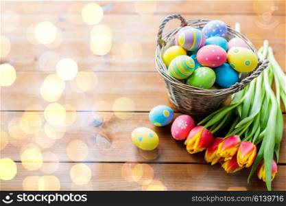 easter, holidays, tradition and object concept - close up of colored easter eggs in basket and tulip flowers on wooden table with copy space over holidays lights