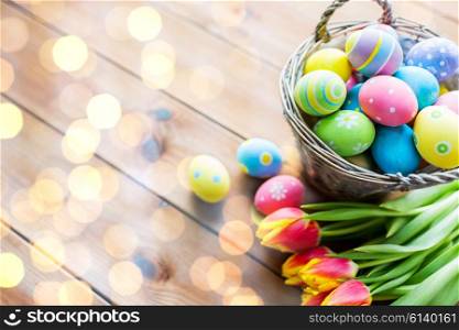 easter, holidays, tradition and object concept - close up of colored easter eggs in basket and tulip flowers on wooden table with copy space over lights