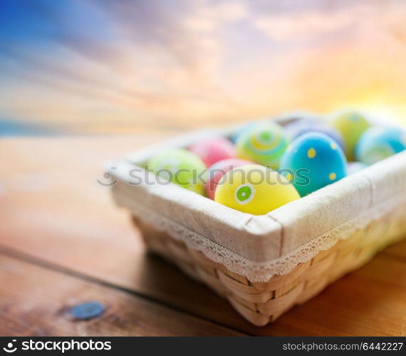 easter, holidays, tradition and object concept - close up of colored eggs in wicker basket on wooden table over sky background. close up of colored easter eggs in basket