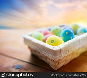 easter, holidays, tradition and object concept - close up of colored eggs in wicker basket on wooden table over sky background. close up of colored easter eggs in basket