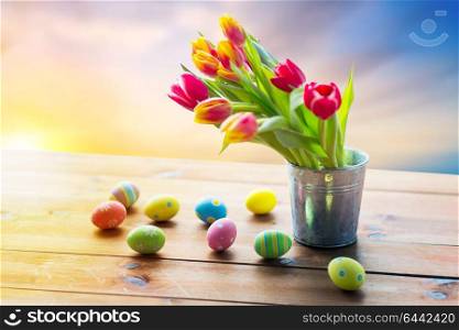 easter, holidays, tradition and object concept - close up of colored eggs and tulip flowers in bucket on wooden table over sky background. close up of easter eggs and flowers in bucket