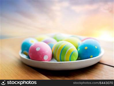easter, holidays, tradition and object concept - close up of colored eggs on plate on wooden table over sky background. close up of colored easter eggs on plate