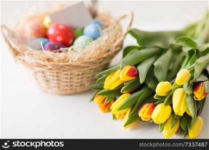 easter, holidays, tradition and object concept - close up of colored eggs in basket and tulip flowers on white background. close up of colored easter eggs and tulip flowers
