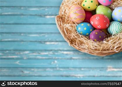 easter, holidays, tradition and object concept - close up of colored eggs in wicker basket over blue wooden boards background. close up of colored easter eggs in basket