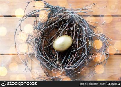 easter, holidays, tradition and object concept - close up of golden easter egg in nest on wooden surface over holidays lights