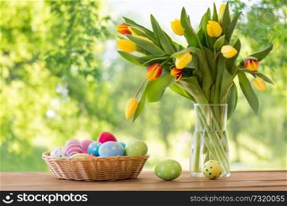 easter, holidays, tradition and object concept - colored eggs in basket and tulip flowers on wooden table over green natural background. colored easter eggs in basket and flowers