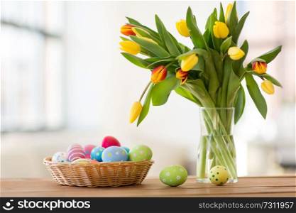 easter, holidays, tradition and object concept - colored eggs in basket and tulip flowers at home. colored easter eggs in basket and flowers at home