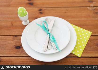 easter, holidays, tradition and object concept - green colored egg in cup holder, plates and cutlery on table at home. easter egg in cup holder, plates and cutlery