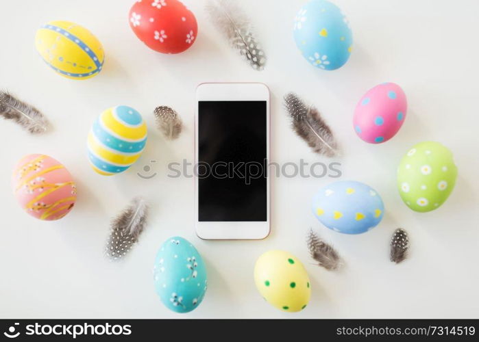 easter, holidays, tradition and object concept - smartphone with colored eggs and quail feathers on white background. smartphone with easter eggs and quail feathers