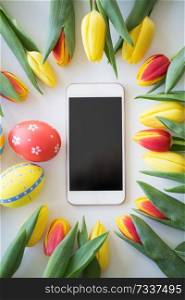 easter, holidays, tradition and object concept - smartphone with colored eggs and tulip flowers on white background. smartphone with easter eggs and tulip flowers