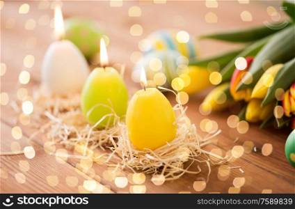 easter, holidays, tradition and object concept - three burning candles in shape of eggs and tulip flowers on wooden table. candles in shape of easter eggs and tulip flowers