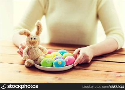 easter, holidays, tradition and people concept - close up of woman hands with colored easter eggs on plate and bunny
