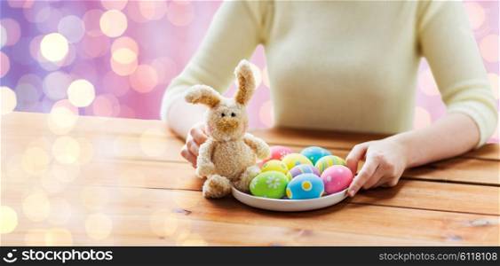 easter, holidays, tradition and people concept - close up of woman hands with colored easter eggs on plate and bunny