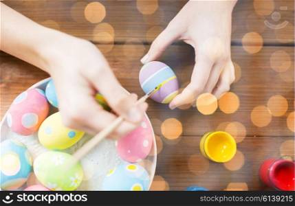 easter, holidays, tradition and people concept - close up of woman hands coloring easter eggs with colors and brush over holidays lights