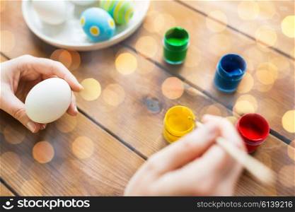 easter, holidays, tradition and people concept - close up of woman hands coloring easter eggs with colors and brush over lights