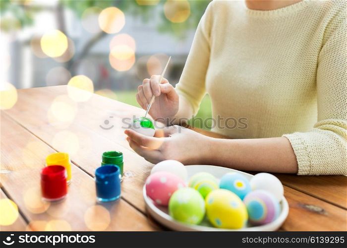 easter, holidays, tradition and people concept - close up of woman hands coloring easter eggs with colors and brush over lights and outdoor background