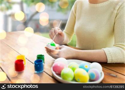 easter, holidays, tradition and people concept - close up of woman hands coloring easter eggs with colors and brush over lights and outdoor background
