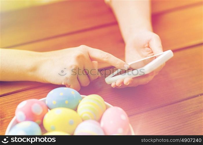 easter, holidays, tradition, technology and people concept - close up of woman hands with colored easter eggs on plate and smartphone. close up of hands with easter eggs and smartphone