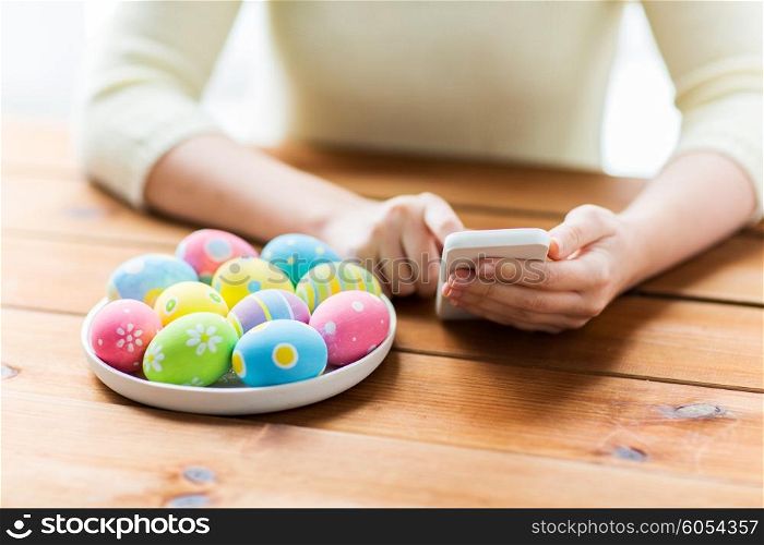 easter, holidays, tradition, technology and people concept - close up of woman hands with colored easter eggs on plate and smartphone