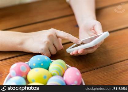 easter, holidays, tradition, technology and people concept - close up of woman hands with colored easter eggs on plate and smartphone