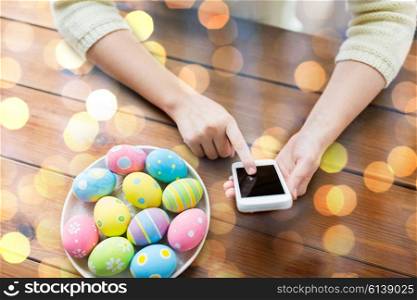 easter, holidays, tradition, technology and people concept - close up of woman hands with colored easter eggs on plate and smartphone over holidays lights