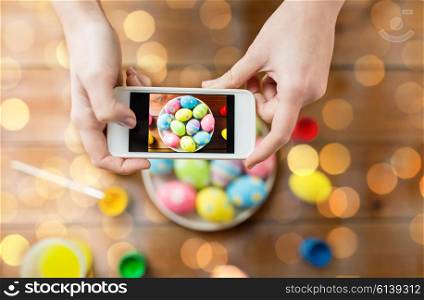 easter, holidays, tradition, technology and people concept - close up of woman hands with smartphone taking picture of colored easter eggs over lights