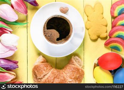 Easter morning with a cup of coffee, rainbow cookies, painted eggs, bunny shaped cookie, croissant and multicolored tulips, on a yellow wooden table.