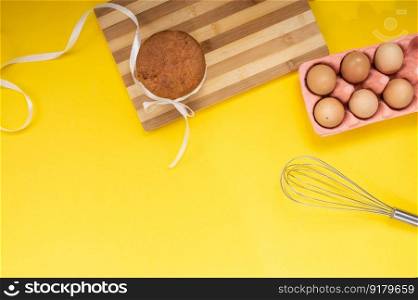 Easter pastries on a wooden board with chicken eggs and a whisk on a yellow background. Top view. Easter pastries on the table with chicken eggs and a whisk on a yellow background. Top view