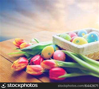 easter, religious holidays and object concept - close up of colored eggs in basket and tulip flowers on wooden table over sky background. close up of easter eggs and flowers on table
