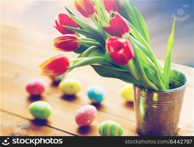easter, religious holidays and object concept - close up of tulip flowers in bucket on wooden table and colored eggs over sky background. tulip flowers in bucket and easter eggs on table
