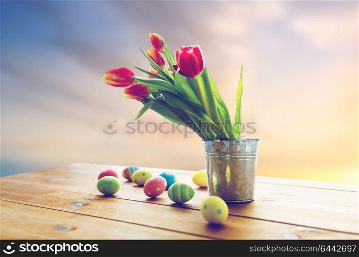 easter, religious holidays and object concept - colored eggs and tulip flowers in bucket on wooden table over sky background. easter eggs and flowers in bucket