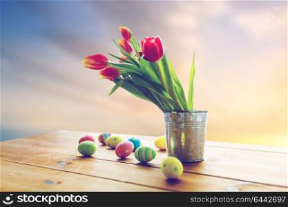 easter, religious holidays and object concept - colored eggs and tulip flowers in bucket on wooden table over sky background. easter eggs and flowers in bucket