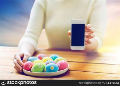 easter, religious holidays, technology and people concept - close up of woman hands with colored eggs on plate and smartphone over sky background. close up of hands with easter eggs and smartphone