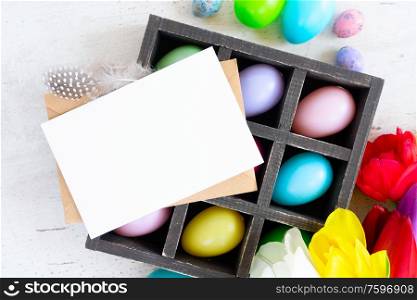 Easter scene with colored eggs in box and tulips, flat lay, copy space on white card. Easter colored eggs with tulips