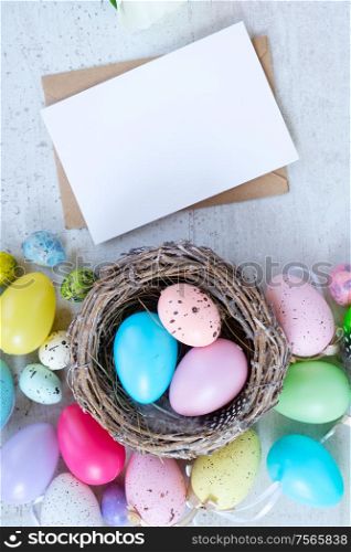 Easter scene with colored eggs in nest, flat lay on white wooden background with copy space on white card. Easter colored eggs