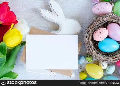 Easter scene with colored eggs, rabbit and tulips, flat lay with copy space on white card. Easter colored eggs with tulips