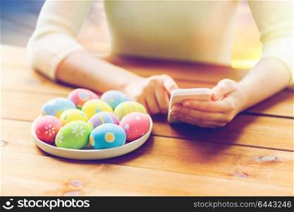easter, technology and people concept - close up of woman hands with colored eggs on plate and smartphone at wooden table. close up of hands with easter eggs and smartphone