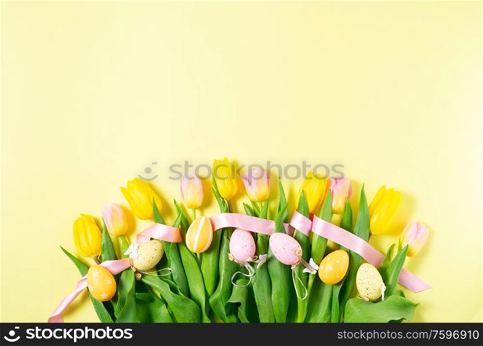 Easter yellow and pink tulip flowers row with colored eggs, top view on yellow background. Easter scene with colored eggs