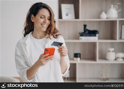 Easy payment with smartphone and card. Happy caucasian girl is going to buy through internet at home. Girl is booking or purchasing online. E-commerce and financial transaction concept.. Easy payment with smartphone and card. Happy caucasian girl is going to buy through internet.