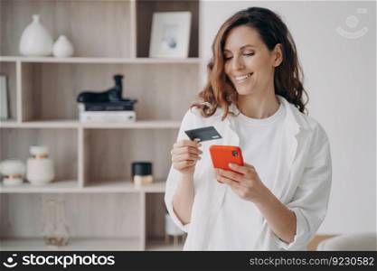 Easy payment with smartphone and card. Happy caucasian girl is going to buy through internet at home. Girl is booking or purchasing online. E-commerce and financial transaction concept.. Easy payment with smartphone and card. Happy caucasian girl is going to buy through internet.