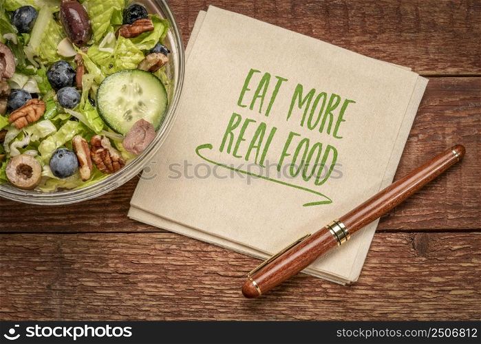 Eat more real food reminder - handwriting on a napkin with a bowl of green salad, healthy eating and lifestyle concept