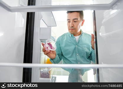 eating and diet concept - middle-aged man taking container with food leftovers from fridge at kitchen. man taking empty food container from fridge