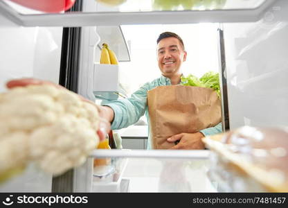 eating, diet and storage concept - smiling middle-aged man with new purchased food in paper bag putting cauliflower to fridge at home. man putting new purchased food to home fridge