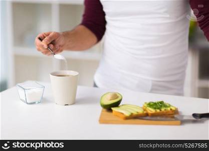 eating, people and diet concept - man adding sugar to tea or coffee cup and having vegetable sandwiches for breakfast at home kitchen. man having breakfast and adding sugar to coffee
