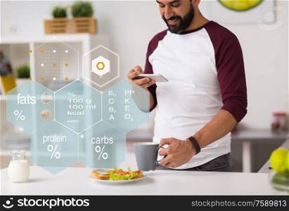 eating, people, technology and diet concept - man with smartphone having vegetable sandwiches and coffee for breakfast at home kitchen over nutritional value chart. man with phone and food nutritional value chart