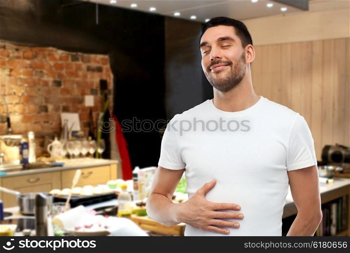 eating, satisfaction and people concept - happy full man touching his tummy over kitchen background