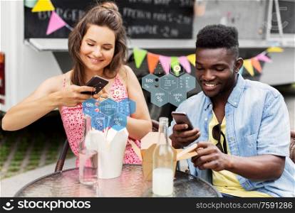 eating, technology and people concept - happy mixed race couple with smartphones and takeaway chinese wok and drinks at food truck over nutritional value chart. couple with smartphones eating wok at food truck