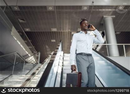 Ebony businessman with briefcase talking by phone on the escalator in mall. Successful business person, black man in formal wear, shopping center