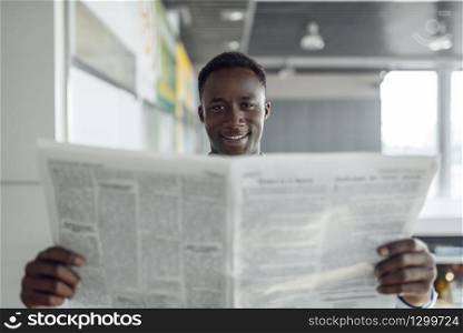 Ebony businessman with newspaper in office hallway. Successful business person reads quotes, black man in formal wear